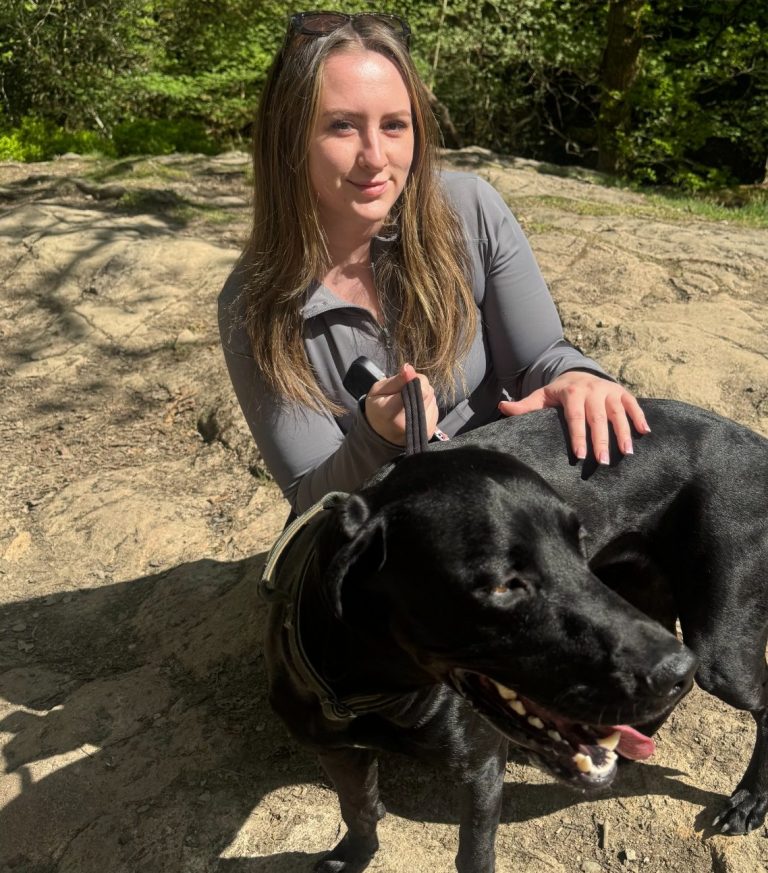 A woman sits on a rock with a black dog in a sunny outdoor setting.